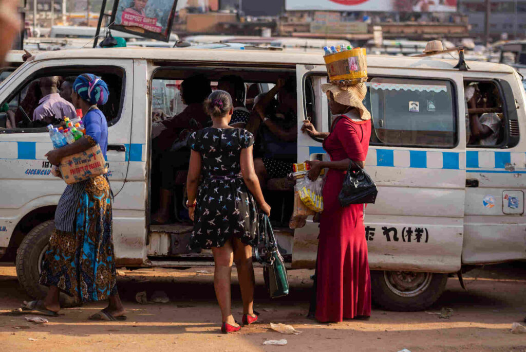 A woman in a long red dress holds a bucket of water bottles on her head, selling to the passengers inside a taxi cab in Kampala, Uganda