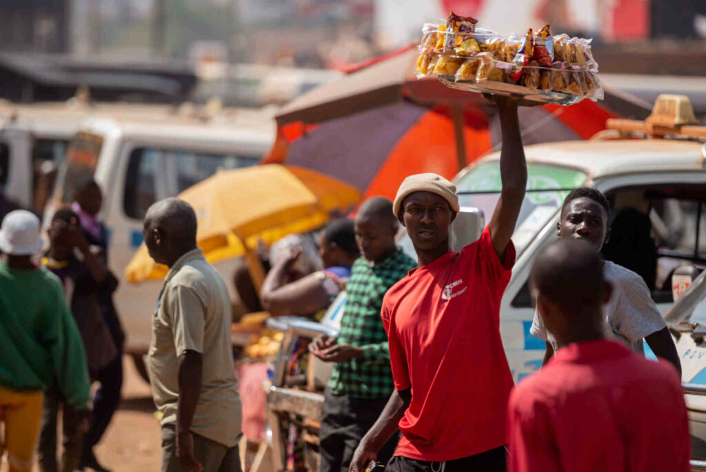 A man wearing a red t-shirt holds a tray of snacks above his head, selling at the Old Taxi Rank in Kampala, Uganda